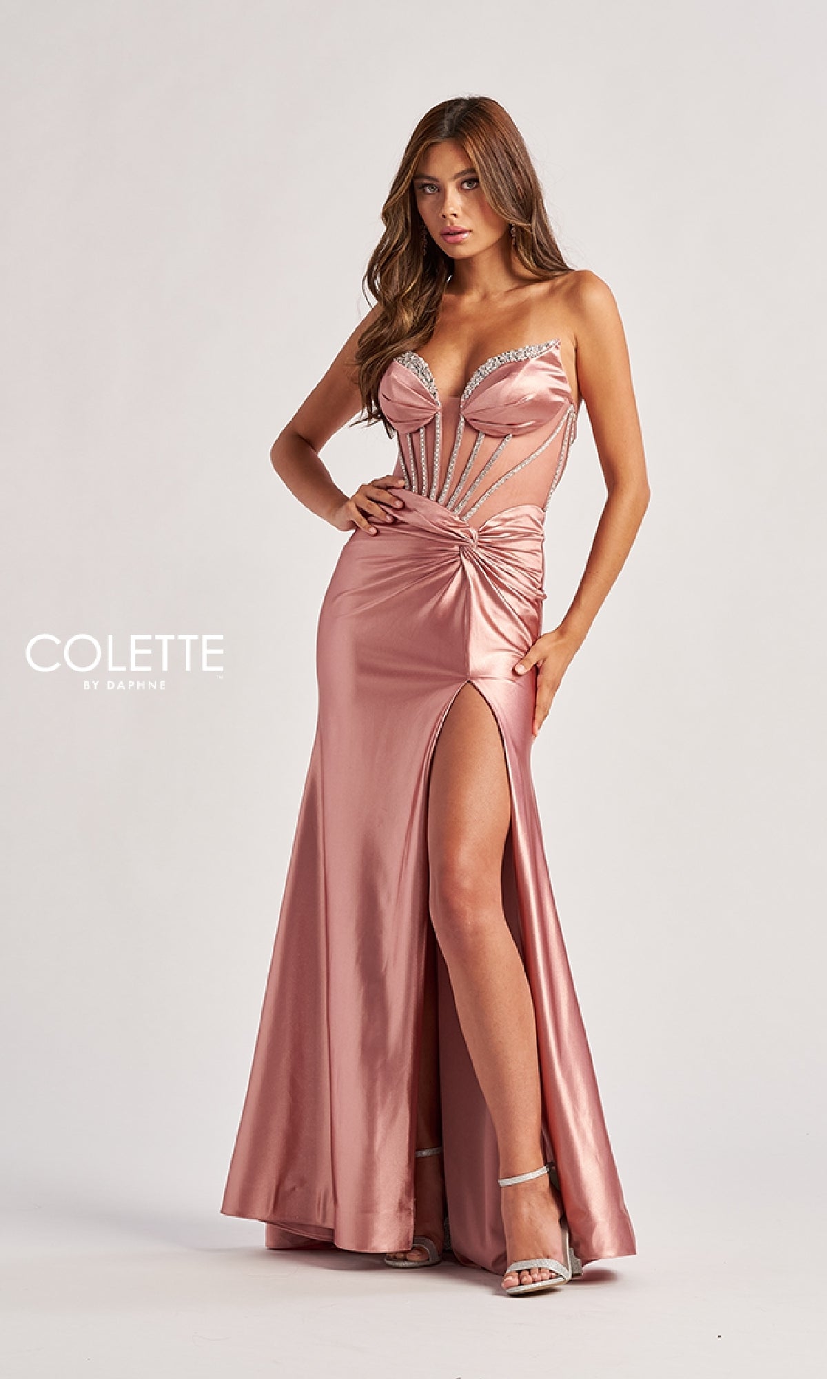 A woman poses against a plain background wearing the Sheer-Bodice Long Prom Dress Colette CL8560, featuring a pink satin strapless design with a thigh-high slit, corset bodice, and silver embellishments.